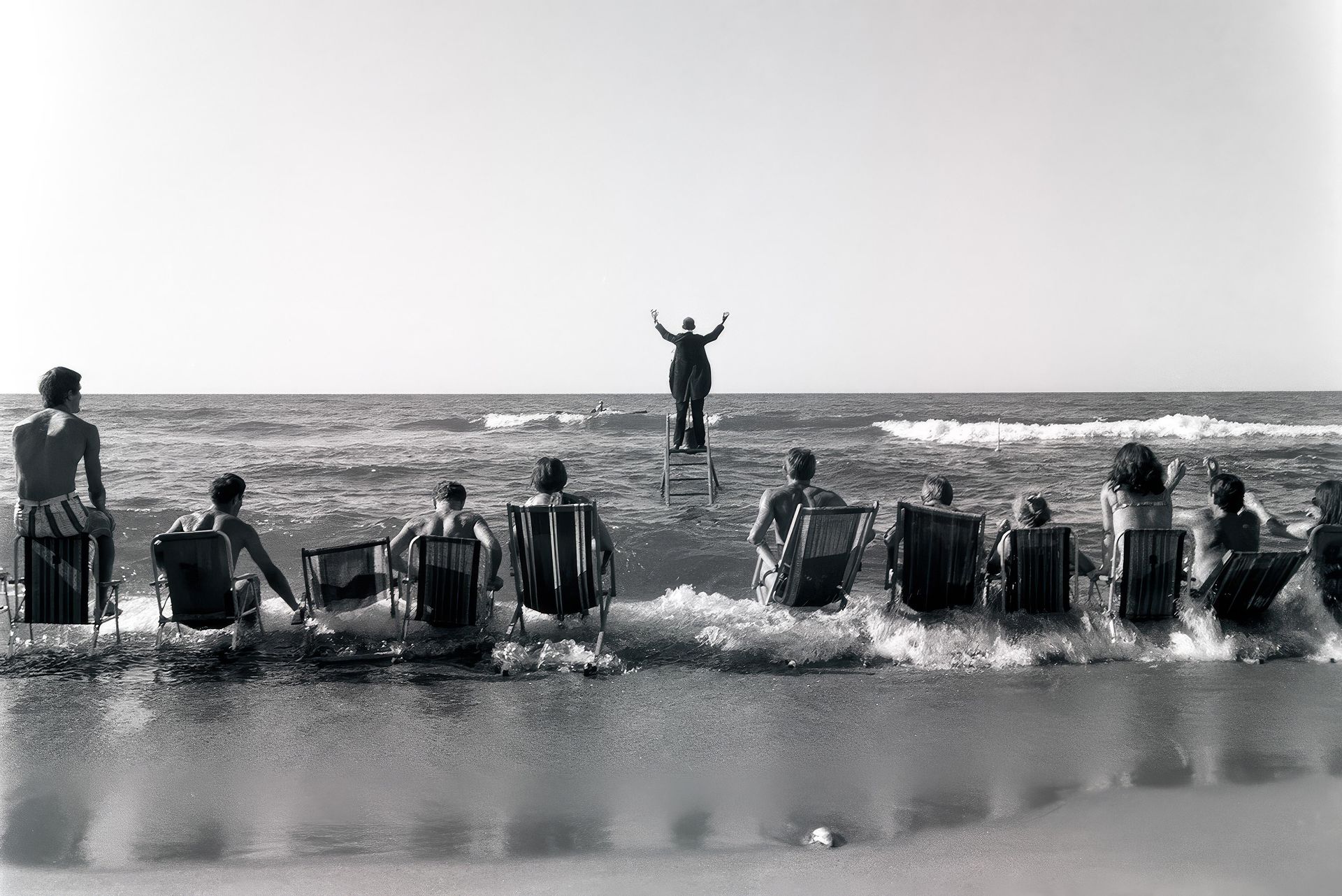 Eine Gruppe von Menschen sitzt in Liegestühlen am Strand, mit Blick auf das Meer. Im Vordergrund stehen die Stühle teilweise im Wasser. Im Hintergrund steht eine Person auf einem Podest oder Stuhl, mit erhobenen Armen, dem Meer zugewandt. Die Szene ist in schwarz-weiß gehalten, das Wasser zeigt leichte Wellen.