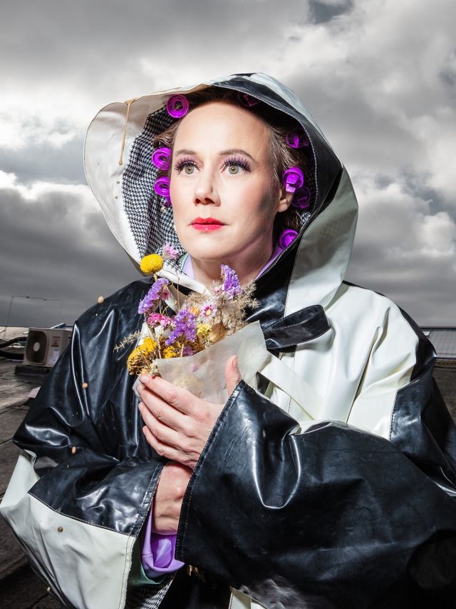 ANTIGONE; Eva Löbau fotographiert von Esra Rotthoff; Die Schauspielerin schaut leicht zur Seite und trägt einen großen, schwarz-weißen Regenmantel. Unter der Kapuze hat sie einige lila Lockenwickler in den Haaren. In den Händen hält sie einen Strauß getrockneter Blumen. Im Hintergrund ist ein bewölkter Himmel zu sehen.