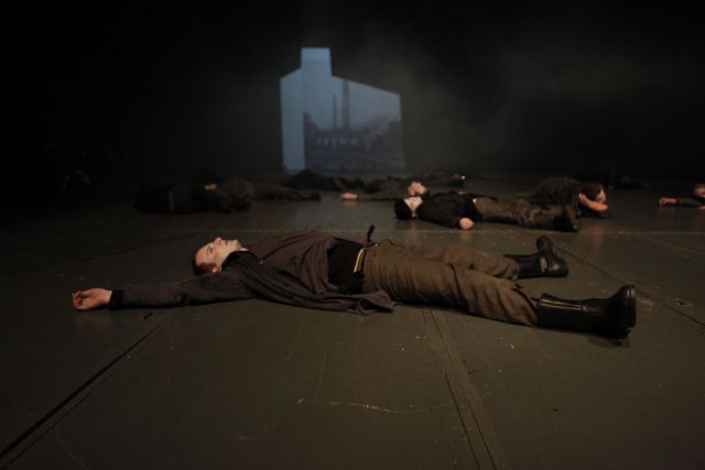 MOJA FABRICA I Schauspieler fotografiert von Muhamed Tunović Badi; 9 Schauspieler liegen ausgestreckt auf ihrem Rücken auf dem Bühnenboden. Man erkennt, dass die Augen des Schauspielers im Vordergrund geöffnet sind. Im Hintergrund sieht man eine schwarz weiß Projektion von einem Haus mit Bäumen.