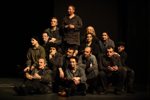 MOJA FABRICA I Schauspieler fotografiert von Muhamed Tunović Badi; 14 Schauspieler sind in einer Pyramiden Form aufgereiht: alle bis auf eine Schauspielerin scheinen zu sitzen. Sie tragen alle dunkle Klamotten, ein Schauspieler hält ein Buch geöffnet. Sie blicken alle mit verschiedenen Gesichtsausdrücken in eine Richtung. Der Hintergrund ist schwarz. 