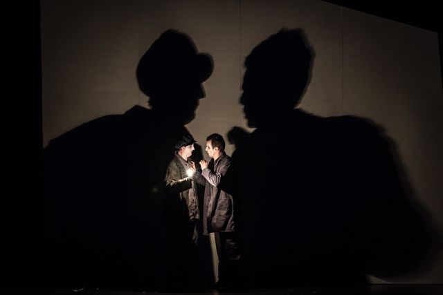 MOJA FABRICA I Schauspieler fotografiert von Veljia Hasanbegović; 2 Schauspieler stehen sich im Dunkeln gegenüber. Sie tragen dunkle Klamotten und haben weiß geschminkte Gesichter mit schwarzen Lippen und Augen. Der linke Mann trägt eine schwarze Mütze und hält eine leuchtende Glühbirne in der Hand. Hinter ihnen erkennt man an der Wand groß abgebildet ihre Schatten.