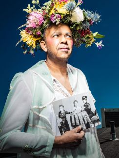 Drei Schwestern (c) Esra Rotthoff Der Schauspieler schaut zur Seite. Er trägt ein weißes Kleid und hält ein schwarz-weißes Bild in der Hand, auf dem drei Frauen zu sehen sind. Auf seinem Kopf befindet sich eine Blumenkrone mit weißen, gelben, blauen und rosa Blumen.