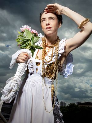 MUTTER COURAGE UND IHRE KINDER; Çiğdem Teke fotografiert von Esra Rotthoff. Die Schauspielerin schaut nach oben. Ihr Haar ist zu einer Hochsteckfrisur gesteckt. Ihre linke Hand liegt auf ihrer Stirn. In ihrer rechten Hand befinden sich rosa Blumen und ein weißes Taschentuch. Sie trägt lange goldene Halsketten, eine weiße Schürze und darunter eine schwarze Jogginghose.