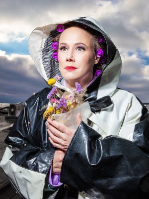 ANTIGONE; Eva Löbau fotographiert von Esra Rotthoff; Die Schauspielerin schaut leicht zur Seite und trägt einen großen, schwarz-weißen Regenmantel. Unter der Kapuze hat sie einige lila Lockenwickler in den Haaren. In den Händen hält sie einen Strauß getrockneter Blumen. Im Hintergrund ist ein bewölkter Himmel zu sehen.